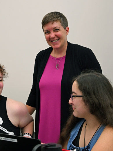 Instructor Tricia Yonker standing between two students talking. She is wearing a black cardigan with a pink shirt underneath and is smiling at the camera. 