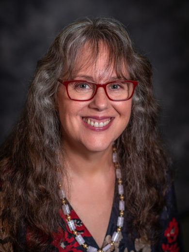 Headshot of MCC instructor Elizabeth Klooster. She is wearing a black and red floral shirt with a large beaded necklace. She has long hair and glasses. 