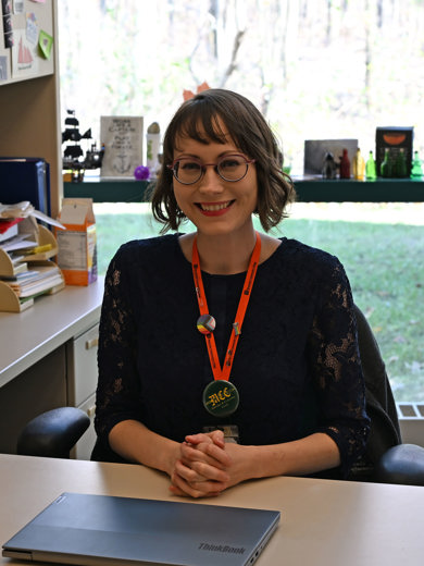 Instructor Andrea Martin sitting at her desk earing a black lace top and smiling at the camera. 