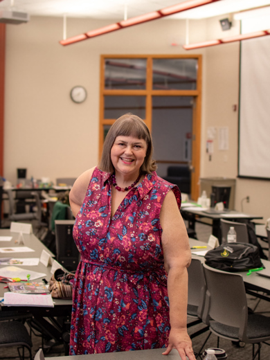 Instructor Greta Skogseth standing in front of a desk waring a pink and blue floral dress and smiling at the camera. 