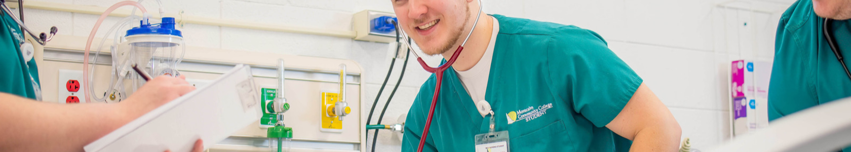 Male nursing student in green scrubs using stethoscope in simulation lab.