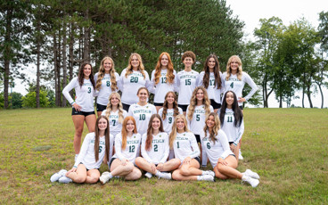 Team shot of the women's volleyball team for the 26-27 season. They are outside sitting on green grass with trees in the background.