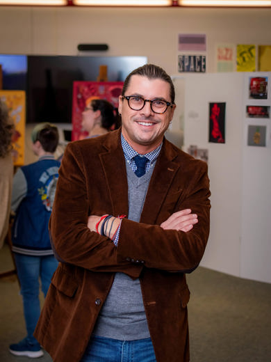 Instructor Seth Sutton standing in a classroom with arms crossed wearing a brown blazer and gray sweater smiling at the camera.