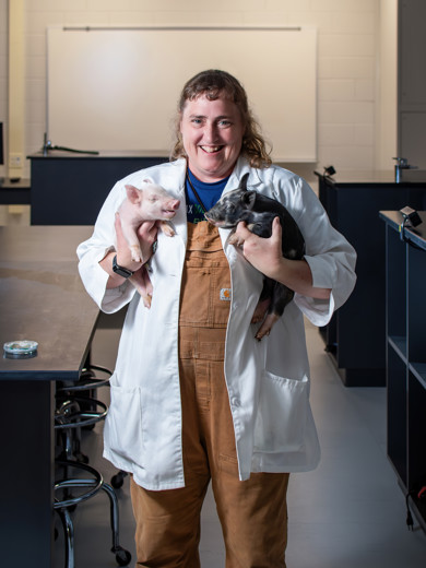Instructor Michelle Gibson standing in a classroom holding a piglet in each arm and smiling at the camera.