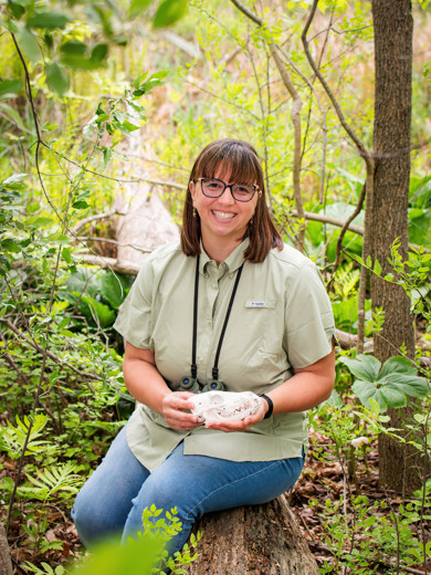 Instructor Heather Wesp sitting on a log outside holding an animal skull and smiling at the camera. 