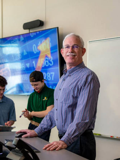 Instructor Bill Bishop standing at a classroom teaching station wearing a blue striped button down shirt and smiling at the camera.