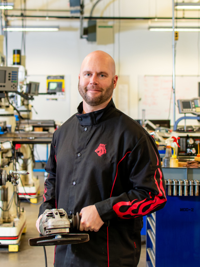 Instructor Andrew Nielsen standing in a classroom holding a power tool and wearing a black welding jacket.