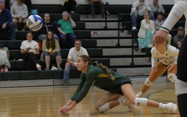 Female volleyball player in green diving to dig the ball during a match with spectators in the background.