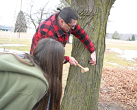 MCC instructor Seth Sutton showing students and community members how to properly tap a maple tree. He is holding a mallet and hitting the tap into the tree.