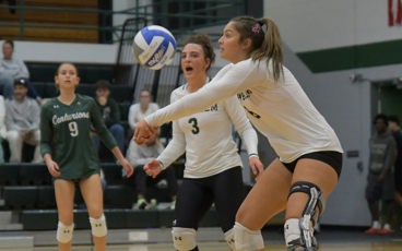 Female volleyball players in white MCC jerseys are preparing to bump the ball during a match indoors.