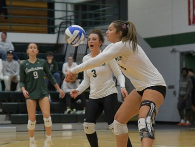 Female volleyball players in white MCC jerseys are preparing to bump the ball during a match indoors.