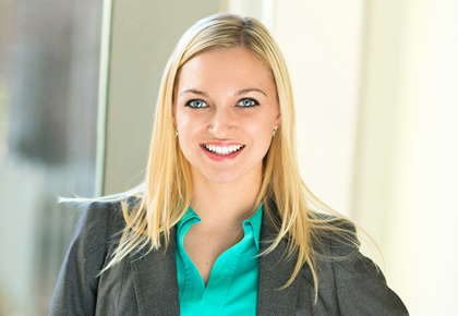 Blonde woman in grey suit with a teal undershirt smiles and looks at camera with her left hand on her hip.