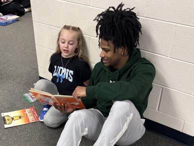 MCC Men's Basketball student volunteering at Central Montcalm Elementary reading with a student on the floor. 