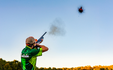 Shooter aiming a shotgun at a clay target in clear blue sky during a shooting competition.