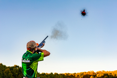 Shooter aiming a shotgun at a clay target in clear blue sky during a shooting competition.