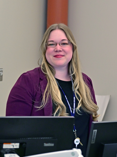 Instructor Devan Bridget standing behind a computer wearing a purple blazer and smiling at the camera.