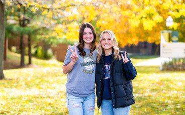 Two female students standing outside on MCC's Sidney campus with yellow fall tress in the background.