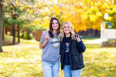 Two female students standing outside on MCC's Sidney campus with yellow fall tress in the background.