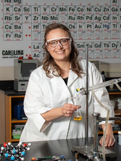 Instrucor Sara Rittersdorf standing in the chemistry lab holding a beaker and smiling at the camera.