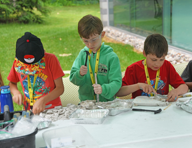 Three students at MCC summer campususing tools to break through plaster to find dinosaurs in dino discovery camp.