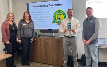 Four people standing together in front of a T.V. screen with text that reads "Ionia County Guarantee"