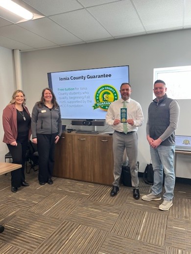 Four people standing together in front of a T.V. screen with text that reads "Ionia County Guarantee"