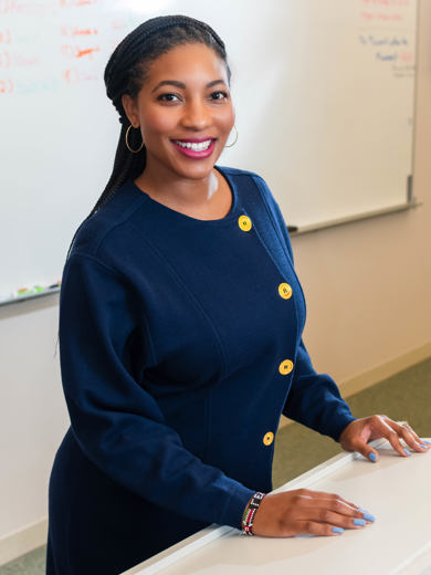 Instructor Leslie Neal standing at a desk wearing a navy blue dress and smiling at the camera.