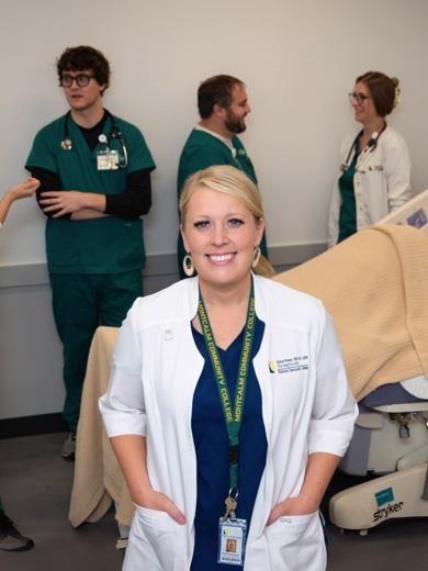 Instrucor Nicole Morris standing in a nursing simulation lab wearing a white coat and scrubs. Students are working with a mannequin in the background.