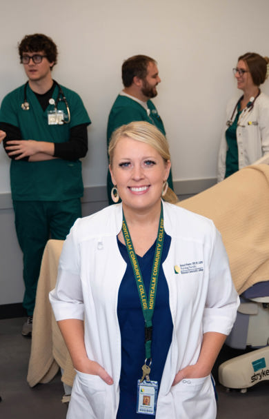 Instrucor Nicole Morris standing in a nursing simulation lab wearing a white coat and scrubs. Students are working with a mannequin in the background.