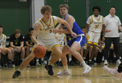 MCC men's basketball player dribbling the ball on the court during a game. An oppossing player attemps to boxout. He is wearing uniform number 5.