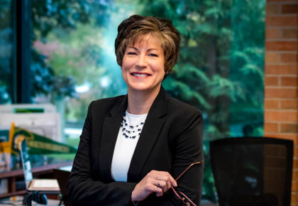 Woman in black suit and white undershirt leans up against brown desk with her arms crossed, holding glasses in her right hand.